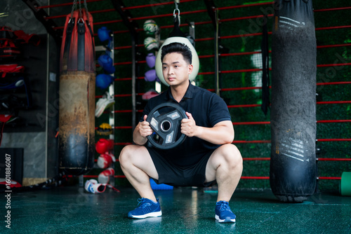 Young Asian man performing a squat exercise with a weight plate in a gym environment focusing on strength training and fitness