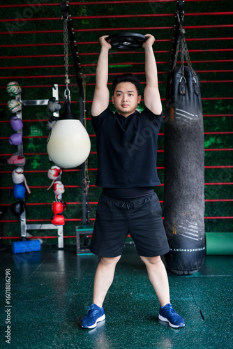 Canvastavla Young Asian man lifting a weight above his head in a fitness gym during a streng
