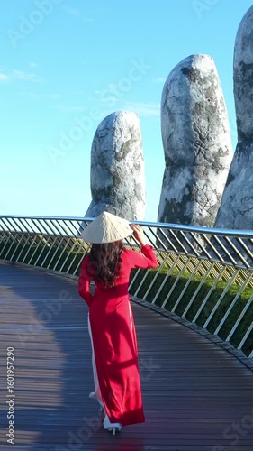 Vertical video of woman wearing red dress, Vietnamese culture with traditional conical hat on Golden Hand Bridge, Da Nang, Vietnam.