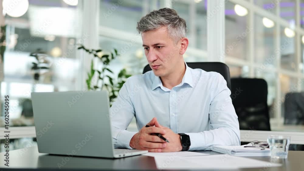 Professional businessman, dressed in a light blue shirt, is focused and engaged during an online video call at a modern office desk, actively discussing important topics. Professional businessman, dressed in a light blue shirt, is focused and engaged during an online video call at a modern office desk, actively discussing important topics.