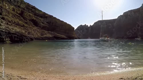 Scenic cove with sandy beach, rocky cliffs, gentle waves and a white sailboat