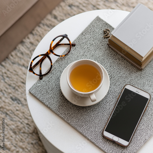 Flat lay of side table with cup of tea, mobile phone, and reading glasses