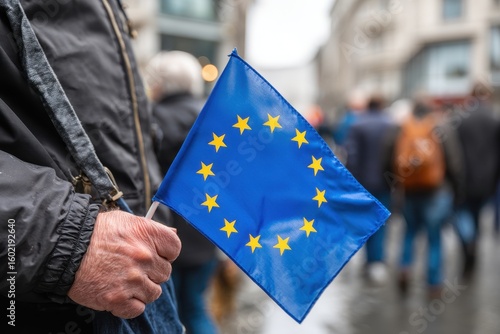 Fototapeta Naklejka Na Ścianę i Meble -  A man in a black jacket holds a small blue EU flag with yellow stars on a daytime street