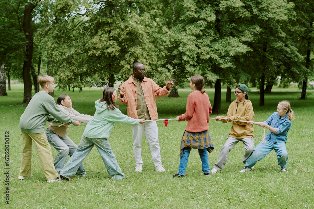 Fototapeta premium Diverse group of children playing tug of war with Black male teacher supervising in park, kids pulling rope and interacting outdoors, teacher encouraging students during activity