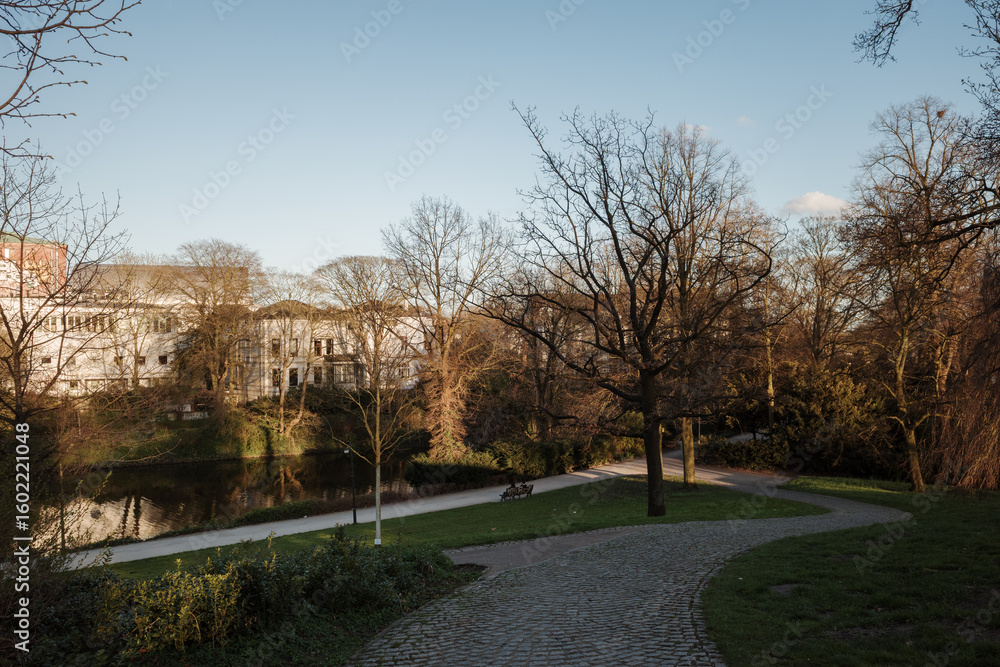 Obraz premium A scenic view of the Wallanlagen park in Bremen, Germany, during early spring or late autumn. A cobblestone path winds through the park next to the historic city moat (Stadtgraben). 