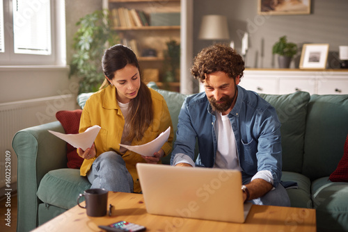 Photography A couple sits together on a couch in their living room, with one partner reviewing life insurance documents while the other works on a laptop