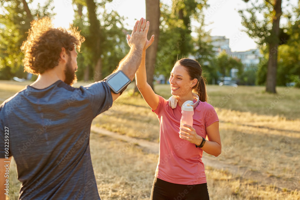 Obraz premium Two individuals enjoy a moment of celebration with a high-five while exercising outdoors in a park. The sun sets behind them, adding warmth to their healthy lifestyle activity.