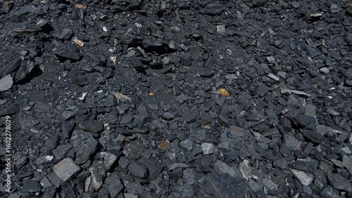 A close-up of dark, coarse gravel and small stones scattered across a rough ground surface, with faint traces of dry leaves and organic debris, suggesting a natural or industrial outdoor setting