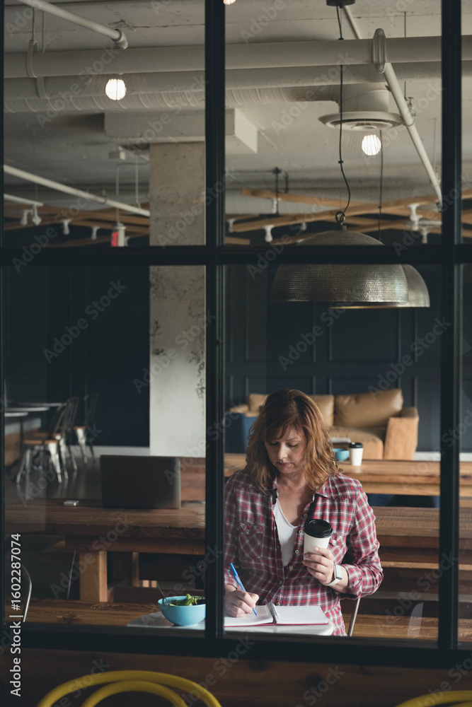 Fototapeta premium Red-haired woman taking notes and holding takeaway coffee cup beside salad bowl at workspace