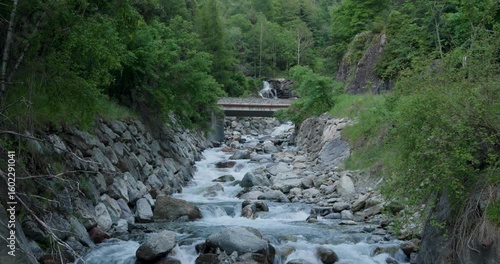 Serene mountain stream flows gently with vibrant greenery and rocky banks near a quiet bridge