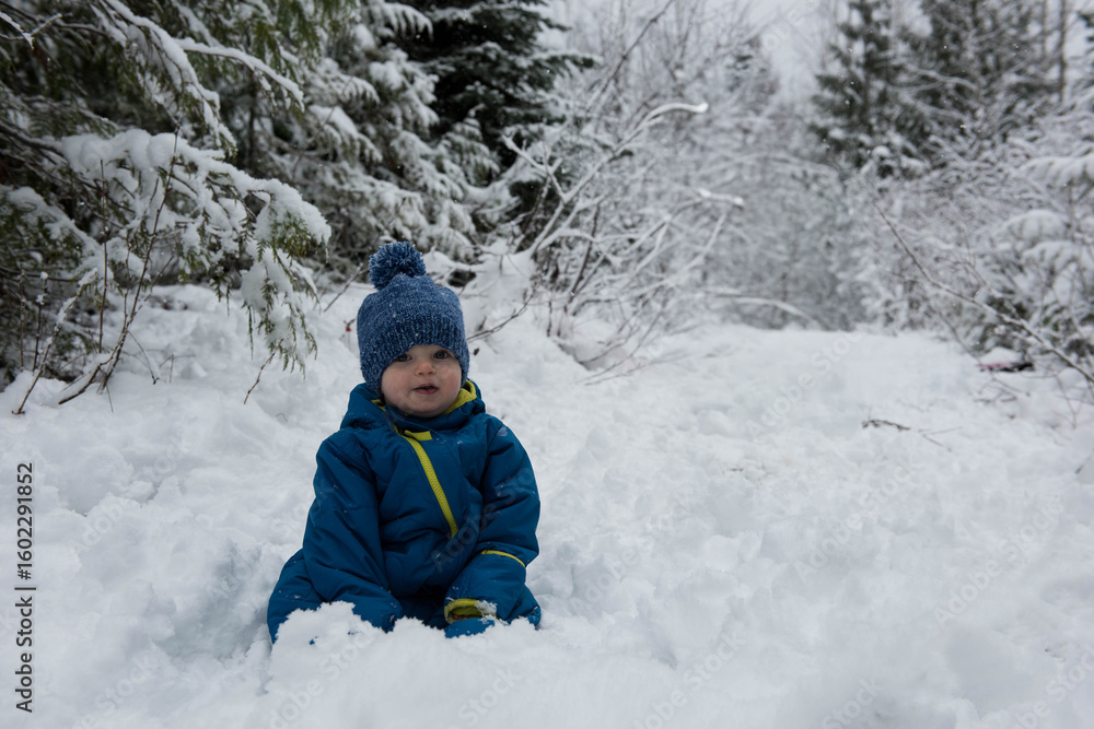 Fototapeta premium Baby boy sitting in deep snow on forest trail wearing blue winter snowsuit and knit hat