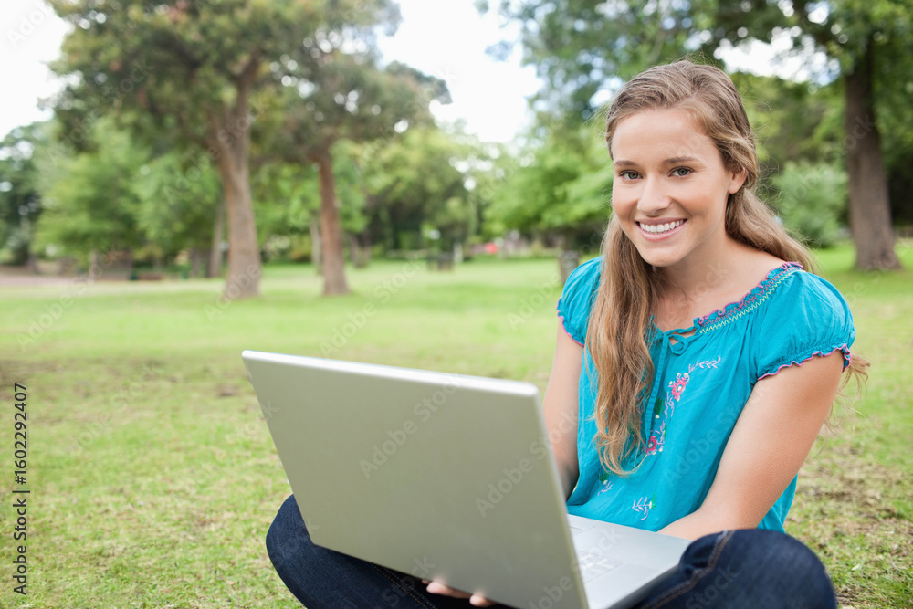 Woman sitting cross-legged on grassy lawn in park using silver laptop, copy space Woman sitting cross-legged on grassy lawn in park using silver laptop, copy space