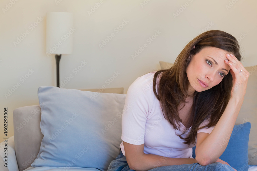 Mid adult woman sitting on sofa holding forehead beside floor lamp in living room, copy space Mid adult woman sitting on sofa holding forehead beside floor lamp in living room, copy space