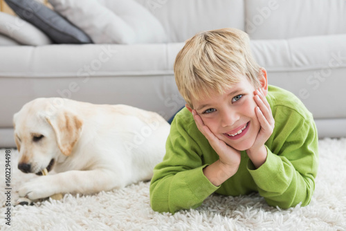 Billede på lærred Blond boy lying on white shag rug at home smiling while puppy chewing bone near
