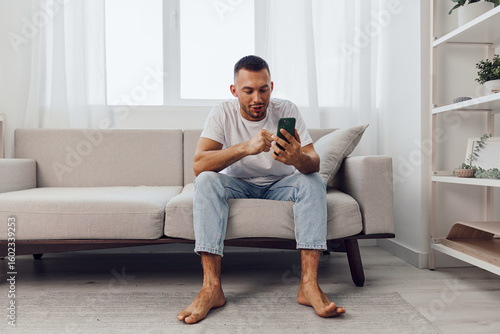 Canvas Print Engaged man sitting on a modern couch, using a smartphone in a bright, minimalistic living room, showcasing the digital lifestyle trend prevalent in 2025-2026