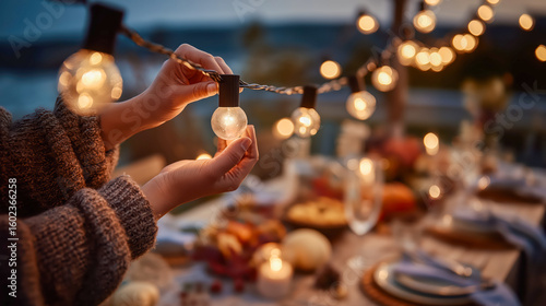 Woman's hands adjusting glowing string light bulbs for a cozy outdoor Thanksgiving day dinner setting, soft warm light
