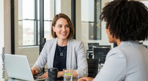 Two women in business attire talking at a desk with a laptop woman meeting