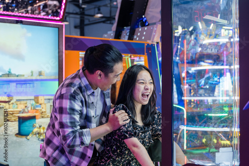 Excited young Asian couple playing an arcade game together in a neon-lit game center, both showing expressive reactions of joy and surprise while looking at the screen in a fun indoor atmosphere