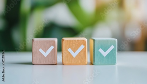 Three pastel-colored wooden blocks, each with a white check mark, sit on a white surface, blurred green plants in the background