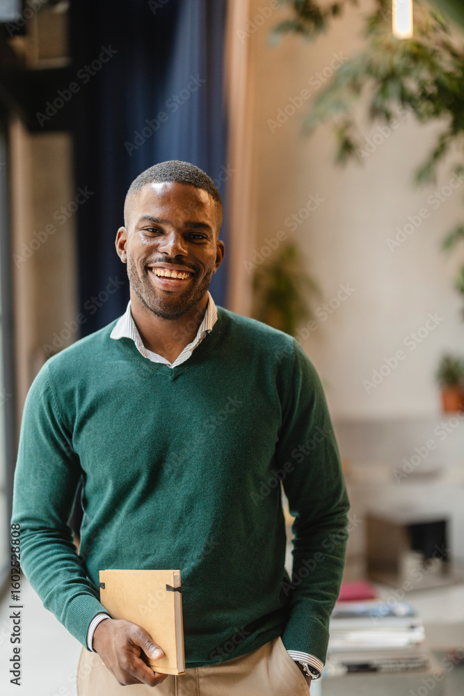 Naklejka premium Smiling black teacher holding notebook in modern classroom