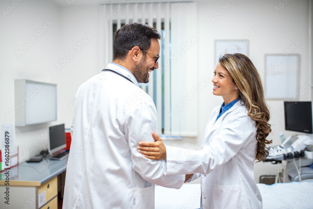 Fototapeta premium Two doctors smile warmly while engaging in a friendly conversation. One places a supportive hand on the other's arm in a gesture of encouragement.