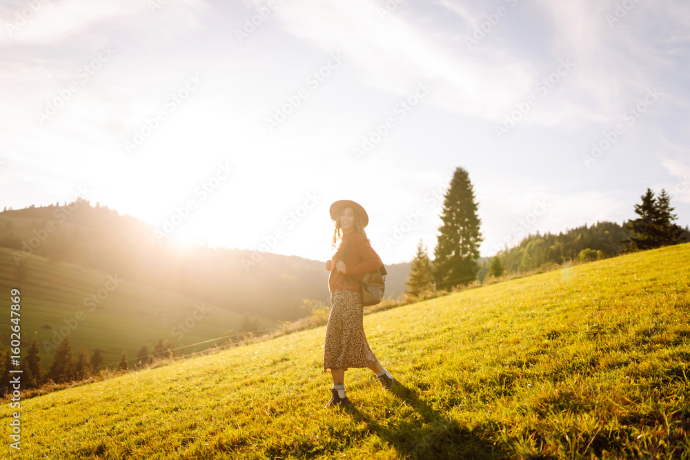 Fototapeta premium Rear view of young female traveler with backpack and hat walking on green hills and enjoying sunset outdoors. Beautiful woman feels freedom against mountain landscape. Hiking and travel concept.
