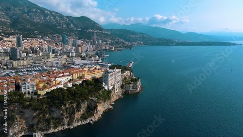 Stunning aerial flight revealing the Oceanographic Museum on the Rock of Monaco, Port Hercules, and the Monte Carlo cityscape against the Mediterranean Sea and mountains.
