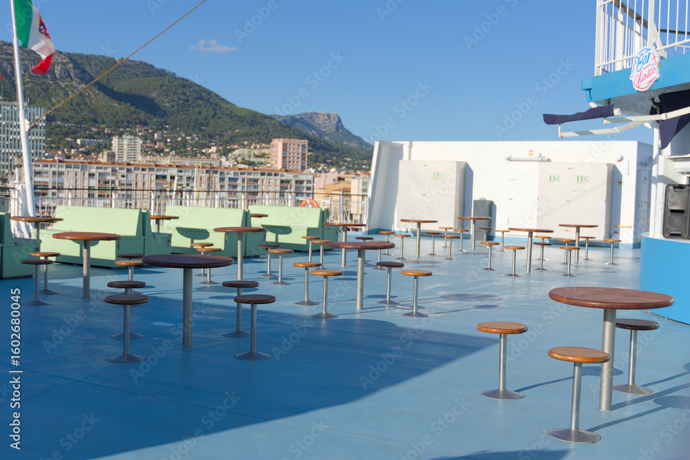 Fototapeta premium An outdoor deck of a ship features numerous high-top tables and stools, green seating, and an Italian flag waving, with a coastal town and mountains in the background under a clear sky.