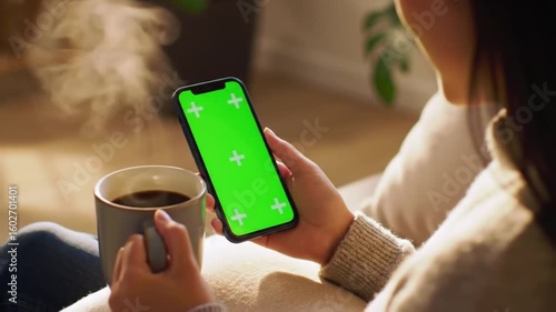 An individual with a smartphone that has a green mock-up screen in upright mode, drinking coffee. The man is navigating the Internet and consuming content, including videos