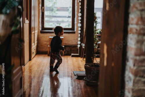 Obraz na plátně A young child walking inside a cozy house with rustic wooden interiors and large windows, creating a warm and inviting ambiance