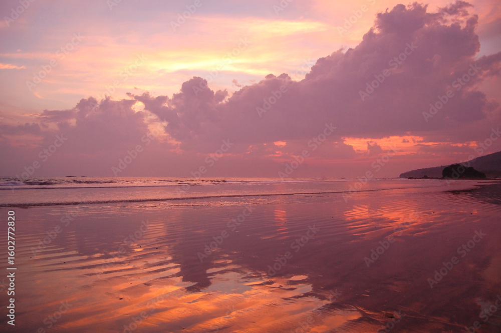 Fototapeta premium A serene beach scene unfolds at sunset, with the sky painted in hues of pink and orange, reflected on the wet sand, creating a tranquil and picturesque moment.