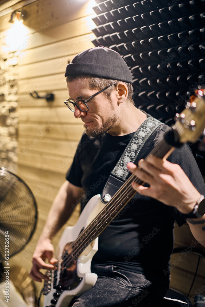Fototapeta premium Young adult male with short hair and glasses plays bass guitar passionately in wooden studio. Warm lighting highlights focused expression. Acoustic panels and fan visible