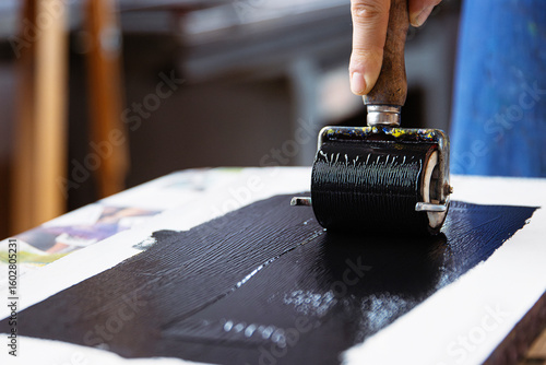 A closeup of an artist hand using a brayer to apply black ink onto a linocut surface in a linocut printmaking process.