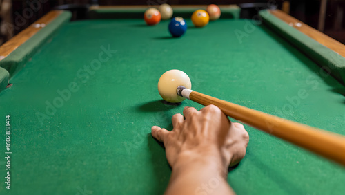 A horizontal image taken from the player's point of view. In the foreground is a hand holding a cue stick about to strike the cue ball (white ball). The cue ball is on a green felt pool table, with th