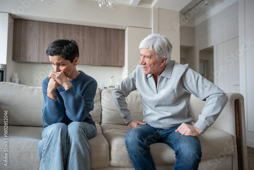 Angry hysterical husband with controlling behavior toward scared wife on couch. Verbal abuse, mental blackmail. Upset mature wife showing fear and withdrawal after argument with manipulative husband