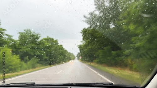 Raindrops on car window focusing side mirror, stormy atmosphere outside, concept of rainy travel, vehicle safety during bad weather. Side mirror reflecting road through rainy car window, overcast sky.
