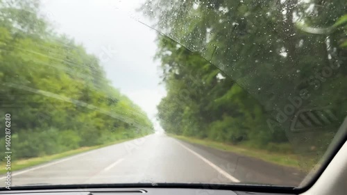 Raindrops on car window focusing side mirror, stormy atmosphere outside, concept of rainy travel, vehicle safety during bad weather. Side mirror reflecting road through rainy car window, overcast sky.