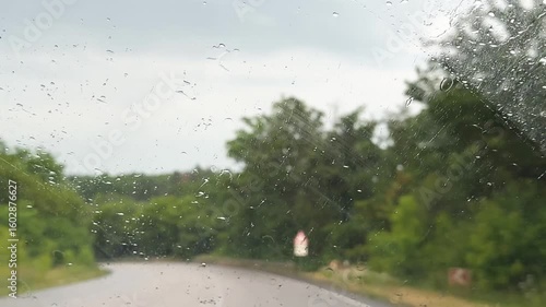 Raindrops on car window focusing side mirror, stormy atmosphere outside, concept of rainy travel, vehicle safety during bad weather. Side mirror reflecting road through rainy car window, overcast sky.