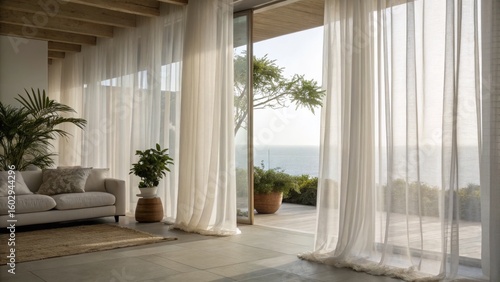 Bright living room with sheer white curtains, indoor plants, and a view of the ocean through large glass doors opening to a wooden deck.