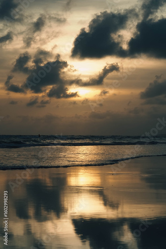 A beautiful sunset over the ocean with the sun's reflection on the waves and wet sand, under a cloudy sky, with distant silhouettes of people in the water.