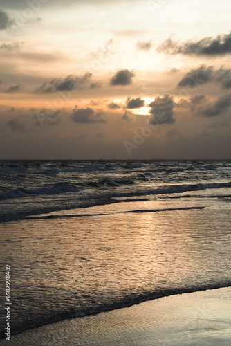 A beautiful sunset over the ocean with the sun's reflection on the waves and wet sand, under a cloudy sky, with distant silhouettes of people in the water.