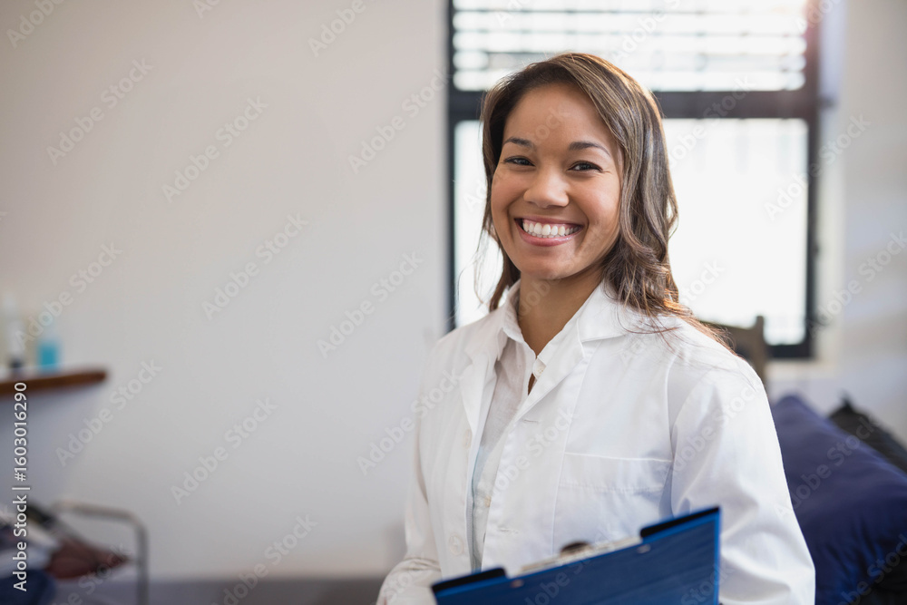 Female healthcare professional smiling and holding blue clipboard in clinical exam room, copy space Female healthcare professional smiling and holding blue clipboard in clinical exam room, copy space