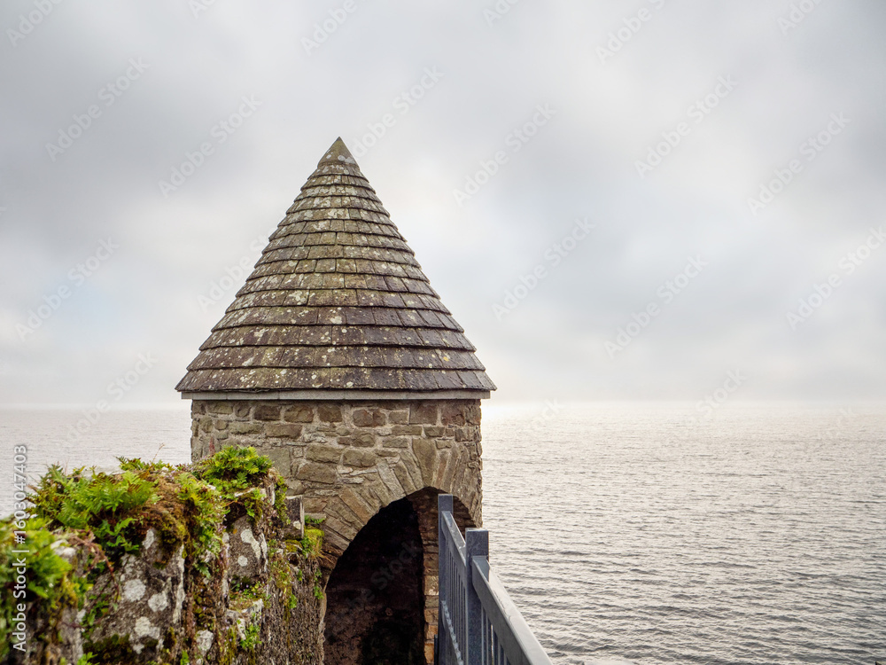 Fototapeta premium A small stone building with a pointed roof sits on a pier overlooking the ocean. The sky is overcast, and the water is calm. The building appears to be a small shelter or castle lookout point
