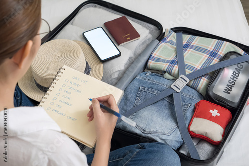 making a check list of things to pack for travel. woman writing checklist item on notebook and packing suitcase to vacation, prepare clothes, medicine, passport into luggage. travel vacation concept