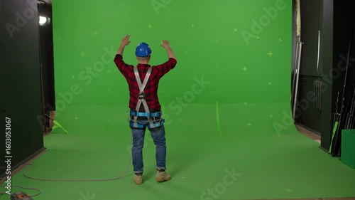 A male builder in professional ammunition and a construction helmet waving his hands and signaling to someone on a green background