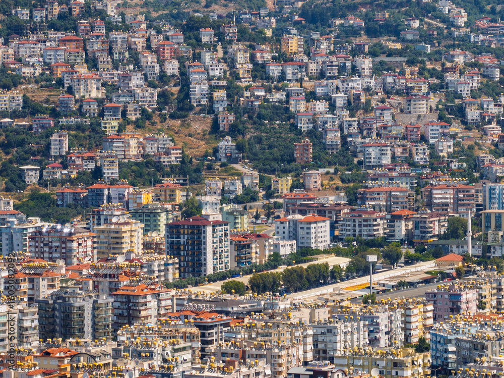 Obraz premium Aerial view of Alanya city in Turkey. Dense urban buildings, Mediterranean coastline, green hills and mountains, from drone during clear summer day. Aerial view of buildings and hotels in Alanya city
