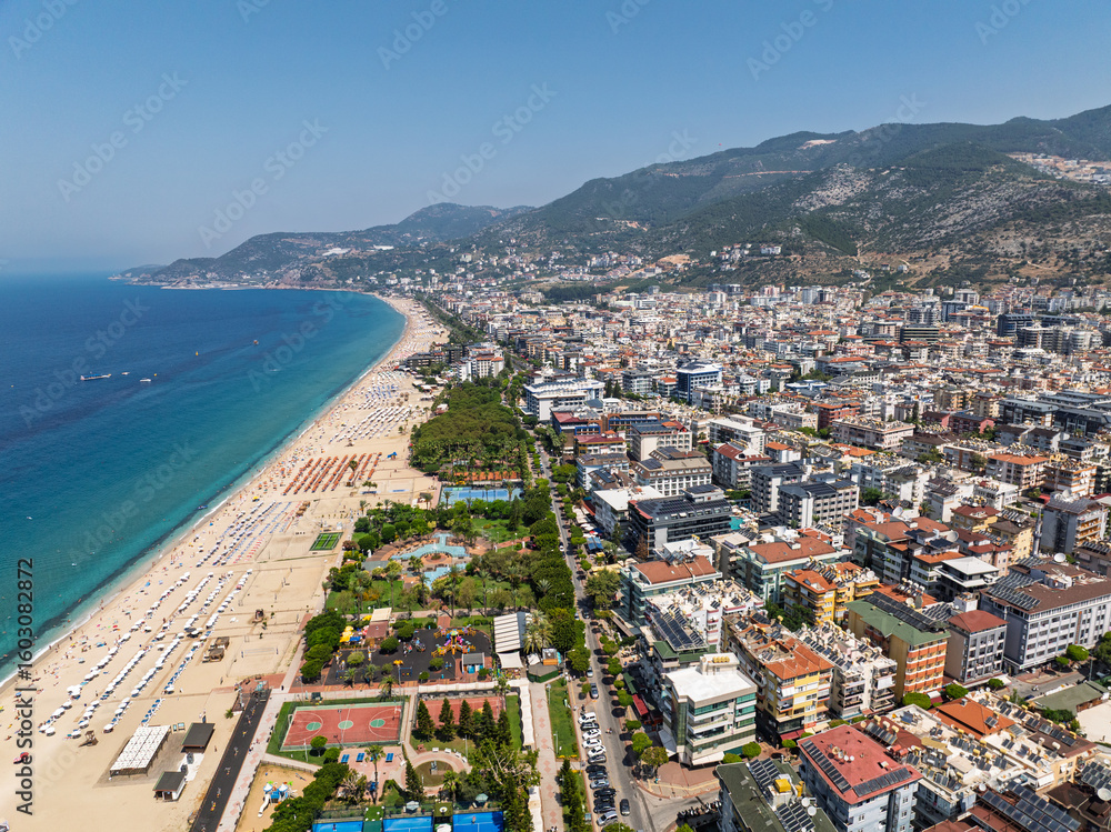 Fototapeta premium Drone view of Alanya city, Turkey. Coastline, Mediterranean Sea, harbor, historic castle on the hill, and sprawling urban area with mountains in the background on a clear summer day. Alanya, Turkey