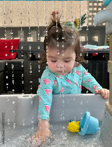 A little girl in her water outfit with her cute pony tail playing on the deck with her water table looking down