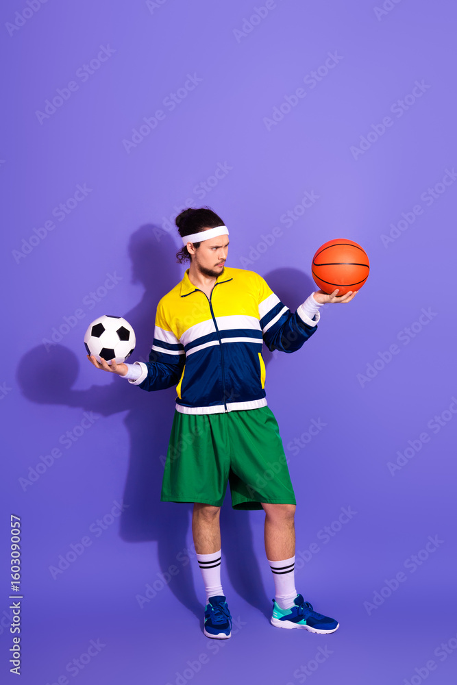 Fototapeta premium Young sporty man in retro-style activewear holding basketball and soccer ball, posing against a vibrant purple backdrop