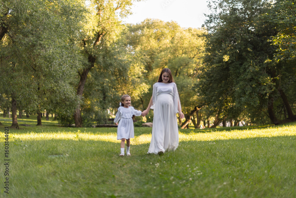 Fototapeta premium A pregnant woman and her daughter are dressed in white clothes and walking in the park. A happy family
